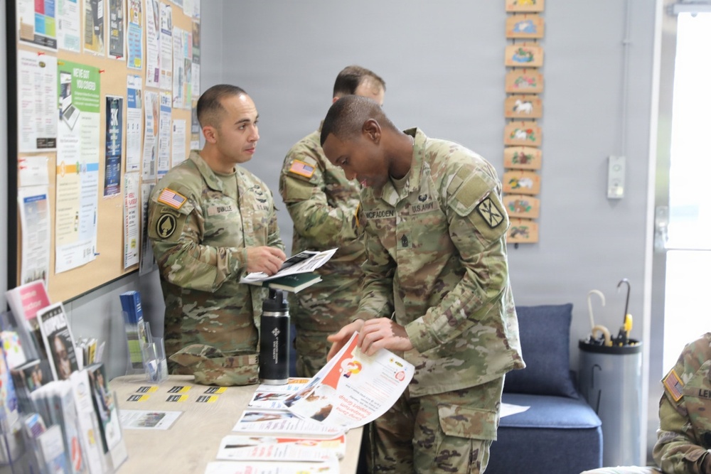 Three U.S. Army soldiers in OCP (Operational Camouflage Pattern) uniforms are gathered near an information table and bulletin board, reviewing printed materials and brochures. The setting appears to be a military resource or outreach center, with pamphlets, flyers, and informational displays visible. The soldiers appear to be engaged in an educational or administrative activity.