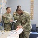 Three U.S. Army soldiers in OCP (Operational Camouflage Pattern) uniforms are gathered near an information table and bulletin board, reviewing printed materials and brochures. The setting appears to be a military resource or outreach center, with pamphlets, flyers, and informational displays visible. The soldiers appear to be engaged in an educational or administrative activity.
