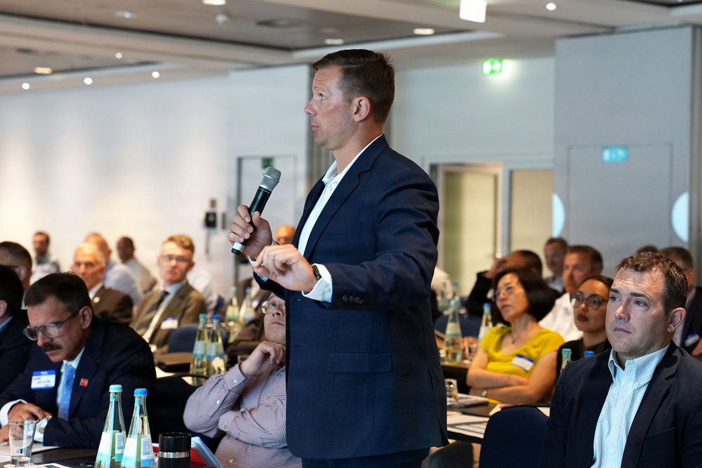 A man in a dark navy blazer stands and speaks into a handheld microphone at what appears to be a professional conference or business meeting. He is surrounded by rows of seated attendees who are listening attentively. The setting is a modern conference room with recessed lighting and glass partitions.
