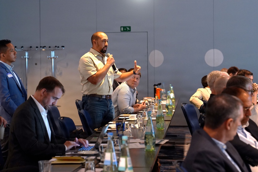 A professional meeting or conference is taking place in a modern conference room. A bald man in a light-colored shirt and jeans is standing and speaking into a microphone while gesturing with one hand. Multiple attendees are seated around a long conference table equipped with water bottles, glasses, and documents.