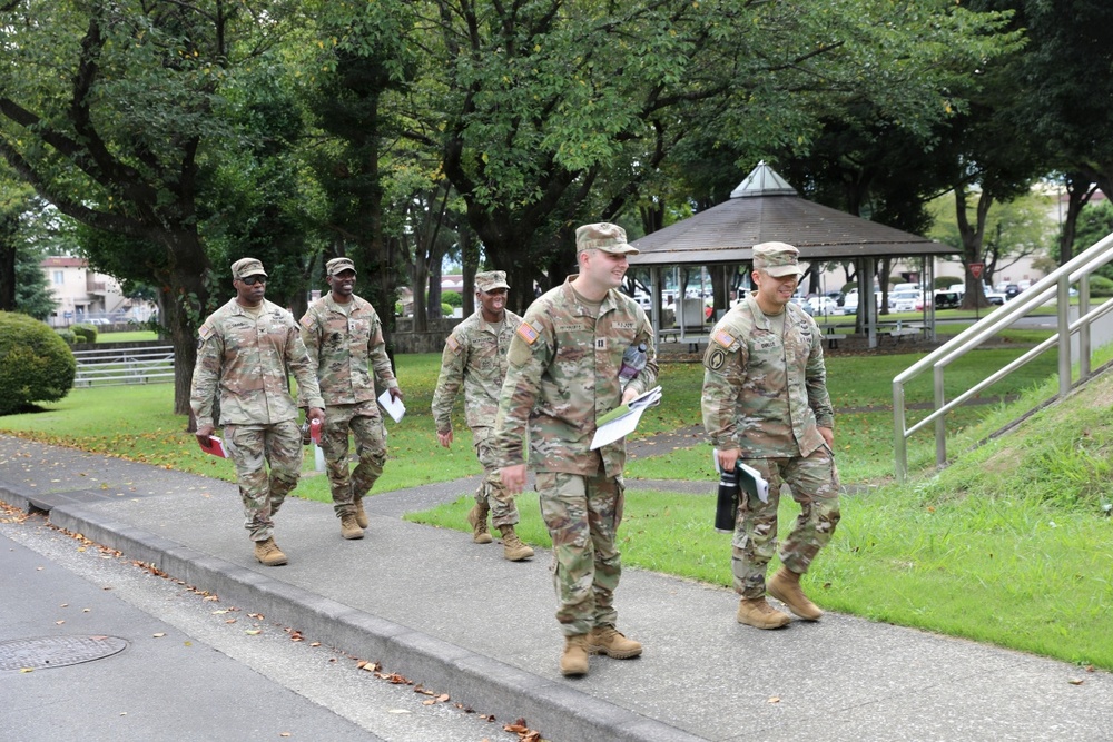 Five U.S. Army soldiers in OCP (Operational Camouflage Pattern) uniforms are walking along a paved path on what appears to be a military installation. They are carrying documents and folders, suggesting an administrative or inspection activity. The setting includes well-maintained grounds with trees, a gazebo structure, and institutional buildings in the background.