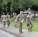 Five U.S. Army soldiers in OCP (Operational Camouflage Pattern) uniforms are walking along a paved path on what appears to be a military installation. They are carrying documents and folders, suggesting an administrative or inspection activity. The setting includes well-maintained grounds with trees, a gazebo structure, and institutional buildings in the background.