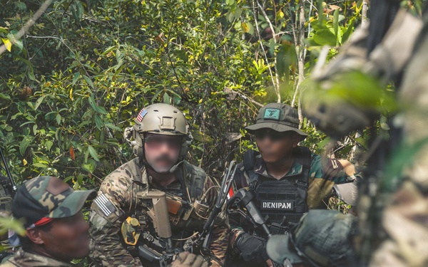 A group of armed military or law enforcement personnel are gathered in a jungle or dense forest environment, appearing to be in a briefing or planning huddle. At least two individuals display US flag patches, and one person wears a vest labeled 'DENIPAM', suggesting a joint multinational operation. Faces have been deliberately blurred, indicating operational security measures.