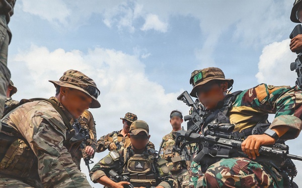 A group of military personnel in mixed camouflage uniforms are gathered in an open sandy field, appearing to conduct a tactical briefing or planning session. One soldier is pointing at what appears to be a map or document on the ground while others crouch or stand around observing. The scene suggests a joint multinational military exercise or training operation.