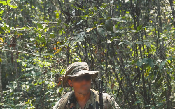 A soldier in full multicam camouflage kit stands on a jungle trail holding an assault rifle with an underbarrel grenade launcher attachment. The individual is equipped with a tactical vest, backpack, boonie hat, and various pieces of kit consistent with a patrol or field exercise. The dense tropical vegetation in the background suggests a jungle or rainforest environment.