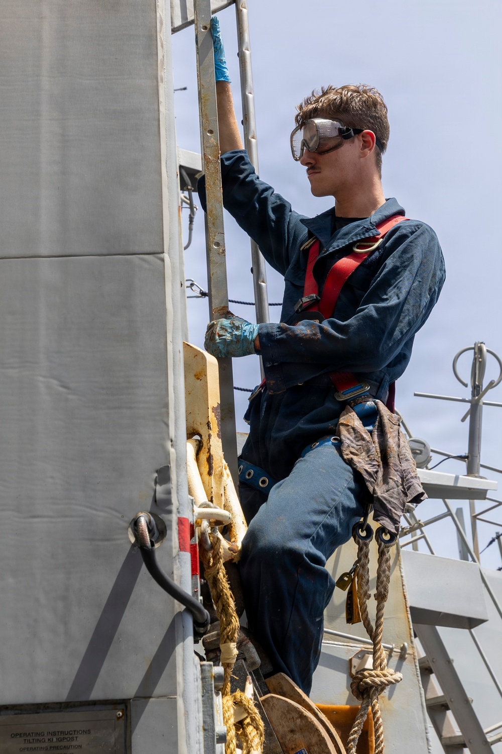 A young worker is climbing a ladder on what appears to be a naval vessel or industrial maritime structure. He is wearing full personal protective equipment including safety goggles, a red safety harness, and blue work gloves while performing maintenance work at height. The environment shows signs of rust and wear consistent with a marine setting.