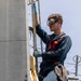 A young worker is climbing a ladder on what appears to be a naval vessel or industrial maritime structure. He is wearing full personal protective equipment including safety goggles, a red safety harness, and blue work gloves while performing maintenance work at height. The environment shows signs of rust and wear consistent with a marine setting.