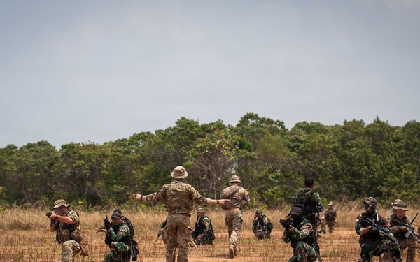 A military training exercise is taking place in an open field with dry grass and sparse vegetation. A central figure in multicam/tan camouflage with a boonie hat appears to be directing or instructing a group of soldiers, some kneeling and some standing, who are armed with rifles. The participants wear mixed camouflage patterns suggesting a multinational or joint training exercise.