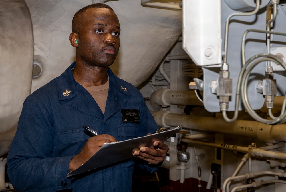 A U.S. Navy enlisted sailor in a dark blue utility uniform is conducting an inspection or maintenance check in what appears to be an engineering or mechanical space aboard a naval vessel. He is holding a clipboard and pen, appearing to record readings or complete a checklist. The background shows various pipes, valves, and mechanical systems typical of a ship's engineering compartment.