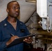 A U.S. Navy enlisted sailor in a dark blue utility uniform is conducting an inspection or maintenance check in what appears to be an engineering or mechanical space aboard a naval vessel. He is holding a clipboard and pen, appearing to record readings or complete a checklist. The background shows various pipes, valves, and mechanical systems typical of a ship's engineering compartment.