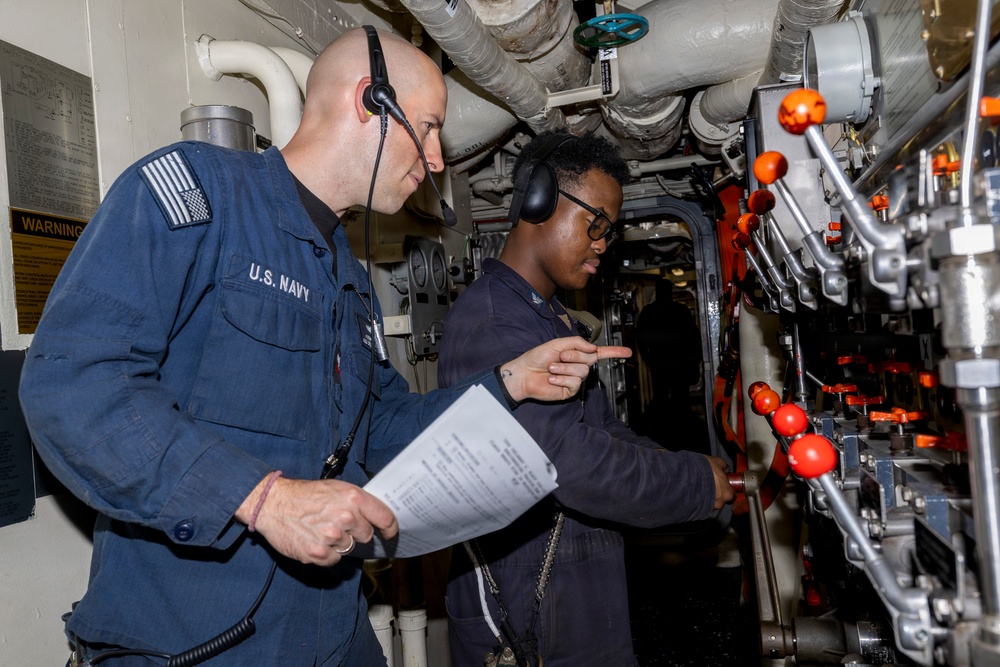 Two U.S. Navy personnel are working together in what appears to be a ship's mechanical or engineering compartment. One sailor wearing a headset holds a document while the other points toward a panel of valves and controls with orange handles. They appear to be conducting a procedure check or inspection using a written checklist.