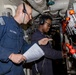 Two U.S. Navy personnel are working together in what appears to be a ship's mechanical or engineering compartment. One sailor wearing a headset holds a document while the other points toward a panel of valves and controls with orange handles. They appear to be conducting a procedure check or inspection using a written checklist.