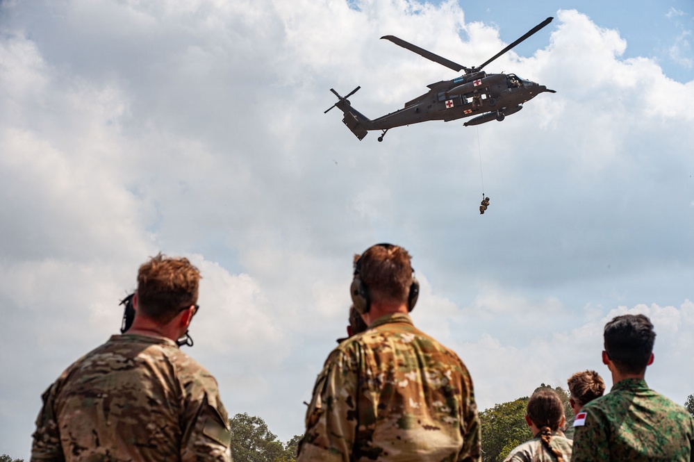 A military medical evacuation (MEDEVAC) helicopter, identifiable by the red cross markings, hovers overhead while conducting a hoist extraction of a person suspended on a cable. Several military personnel in camouflage uniforms observe the operation from the ground, including soldiers in what appears to be US Army OCP pattern and at least one soldier in a different camouflage pattern with a visible red and white flag patch. The scene appears to be a training exercise or demonstration.