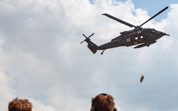 A military medical evacuation (MEDEVAC) helicopter, identifiable by the red cross markings, hovers overhead while conducting a hoist extraction of a person suspended on a cable. Several military personnel in camouflage uniforms observe the operation from the ground, including soldiers in what appears to be US Army OCP pattern and at least one soldier in a different camouflage pattern with a visible red and white flag patch. The scene appears to be a training exercise or demonstration.