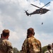 A military medical evacuation (MEDEVAC) helicopter, identifiable by the red cross markings, hovers overhead while conducting a hoist extraction of a person suspended on a cable. Several military personnel in camouflage uniforms observe the operation from the ground, including soldiers in what appears to be US Army OCP pattern and at least one soldier in a different camouflage pattern with a visible red and white flag patch. The scene appears to be a training exercise or demonstration.