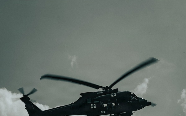 A military medical helicopter is performing a hoist rescue operation, with a person suspended on a cable beneath the aircraft. The helicopter displays white cross markings indicating its medical/medevac designation. The image is captured from a low angle against a cloudy sky in a desaturated, high-contrast tone.