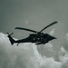 A military medical helicopter is performing a hoist rescue operation, with a person suspended on a cable beneath the aircraft. The helicopter displays white cross markings indicating its medical/medevac designation. The image is captured from a low angle against a cloudy sky in a desaturated, high-contrast tone.