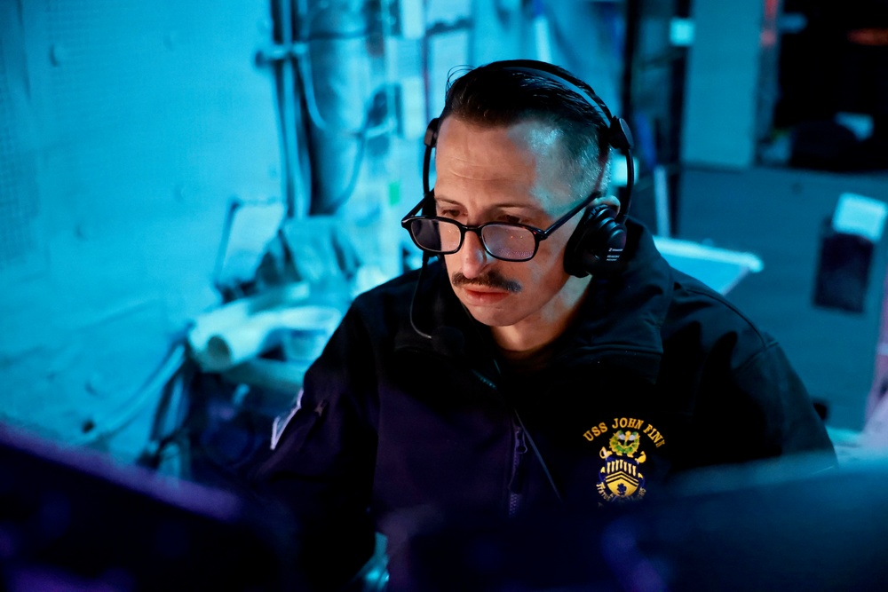 A naval personnel member is seated at a workstation in what appears to be a dimly lit operations or combat information center aboard a naval vessel. The individual is wearing a black jacket bearing the 'USS John Fin' insignia with a chief petty officer rank emblem, along with headphones and glasses, and appears to be monitoring equipment. The scene is illuminated predominantly by blue ambient lighting, typical of shipboard tactical spaces.