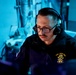 A naval personnel member is seated at a workstation in what appears to be a dimly lit operations or combat information center aboard a naval vessel. The individual is wearing a black jacket bearing the 'USS John Fin' insignia with a chief petty officer rank emblem, along with headphones and glasses, and appears to be monitoring equipment. The scene is illuminated predominantly by blue ambient lighting, typical of shipboard tactical spaces.