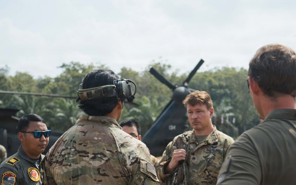 A group of military personnel from what appears to be multiple nations are gathered in an outdoor briefing or discussion on a field area. At least one individual wears a US Army patch and another wears what appears to be an Indonesian Air Force flight suit with squadron patches. A military helicopter is visible in the background.