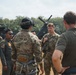 A group of military personnel from what appears to be multiple nations are gathered in an outdoor briefing or discussion on a field area. At least one individual wears a US Army patch and another wears what appears to be an Indonesian Air Force flight suit with squadron patches. A military helicopter is visible in the background.