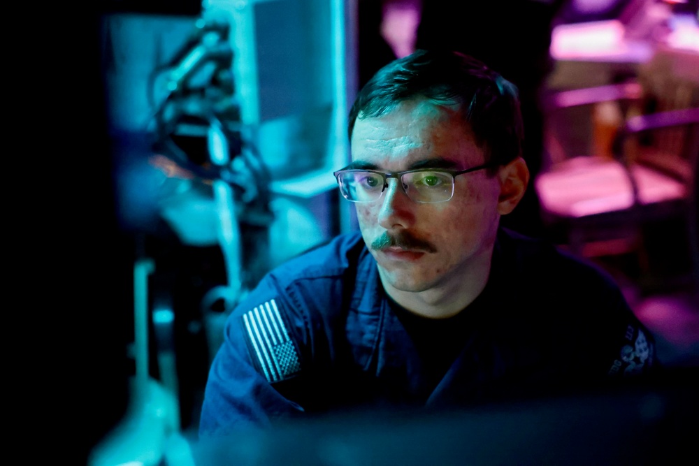A young male in a dark blue military uniform with a US flag patch on the left shoulder is seated and focused on a screen in a dimly lit environment. The scene is illuminated by blue and purple ambient lighting, suggesting a tactical operations or command center setting. The subject appears to be actively monitoring or working at a workstation.