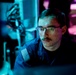 A young male in a dark blue military uniform with a US flag patch on the left shoulder is seated and focused on a screen in a dimly lit environment. The scene is illuminated by blue and purple ambient lighting, suggesting a tactical operations or command center setting. The subject appears to be actively monitoring or working at a workstation.