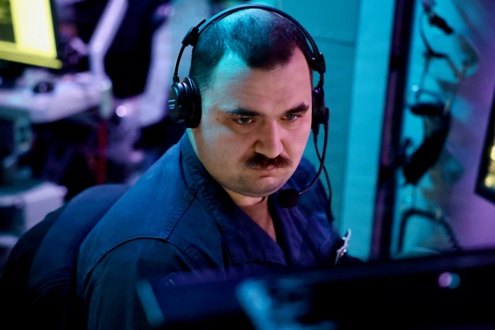 A man wearing a black communications headset with a boom microphone is focused intently on equipment in front of him. He is dressed in a dark blue uniform or work shirt and has a mustache. The environment is dimly lit with blue and purple ambient lighting, suggesting a control room, operations center, or technical workspace.