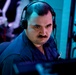 A man wearing a black communications headset with a boom microphone is focused intently on equipment in front of him. He is dressed in a dark blue uniform or work shirt and has a mustache. The environment is dimly lit with blue and purple ambient lighting, suggesting a control room, operations center, or technical workspace.
