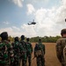 A group of military personnel from at least two different nations are observing a helicopter hoist or rappel operation in an open field. A dark military helicopter, likely a UH-60 Black Hawk, is hovering at altitude with a person suspended beneath it. The scene appears to be a joint military training exercise conducted in a tropical environment.