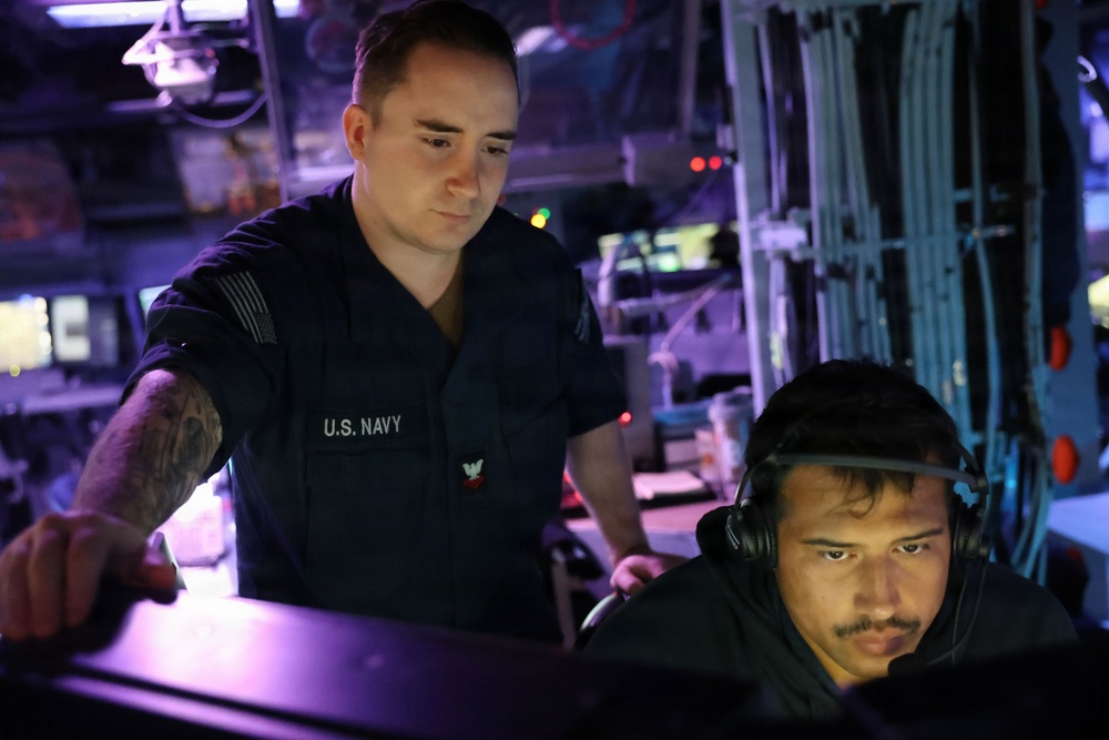 Two U.S. Navy personnel are working at what appears to be a combat information center or operations room aboard a naval vessel. One sailor in a U.S. Navy uniform is standing and pointing at a monitor, while a second sailor wearing headphones is seated and focused on the screen. The environment is dimly lit with blue and purple ambient lighting typical of shipboard operations spaces.