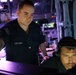 Two U.S. Navy personnel are working at what appears to be a combat information center or operations room aboard a naval vessel. One sailor in a U.S. Navy uniform is standing and pointing at a monitor, while a second sailor wearing headphones is seated and focused on the screen. The environment is dimly lit with blue and purple ambient lighting typical of shipboard operations spaces.
