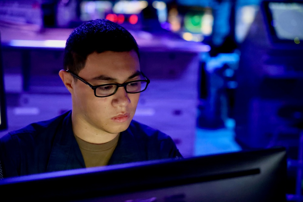 A young person wearing black-framed glasses is focused on a computer monitor in a dimly lit, blue-purple illuminated environment. The setting appears to be a technical operations room or command center with multiple screens and equipment visible in the background. The subject is wearing a dark jacket over a tan/olive undershirt and appears deeply concentrated on the screen in front of them.