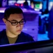 A young person wearing black-framed glasses is focused on a computer monitor in a dimly lit, blue-purple illuminated environment. The setting appears to be a technical operations room or command center with multiple screens and equipment visible in the background. The subject is wearing a dark jacket over a tan/olive undershirt and appears deeply concentrated on the screen in front of them.