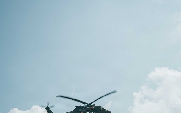 A military medical evacuation (MEDEVAC) helicopter, marked with red cross symbols, hovers over an open field while hoisting a person via a cable/hoist system. Two personnel are visible on the ground below, appearing to observe or assist with the operation. The scene takes place in a tropical environment with dense tree cover in the background.