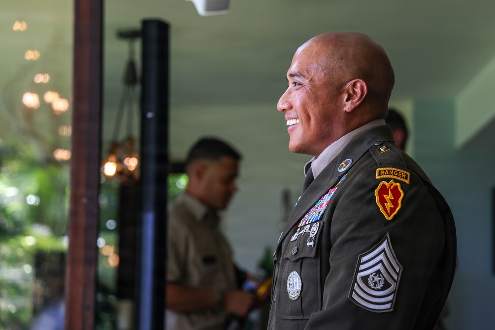 A smiling U.S. Army Sergeant Major in dress green uniform is photographed in profile, displaying multiple medals, unit patches, and rank insignia. He wears a Ranger tab and a 25th Infantry Division shoulder sleeve insignia, indicating significant military service and qualifications. A second military individual is visible in soft focus in the background.