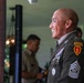 A smiling U.S. Army Sergeant Major in dress green uniform is photographed in profile, displaying multiple medals, unit patches, and rank insignia. He wears a Ranger tab and a 25th Infantry Division shoulder sleeve insignia, indicating significant military service and qualifications. A second military individual is visible in soft focus in the background.