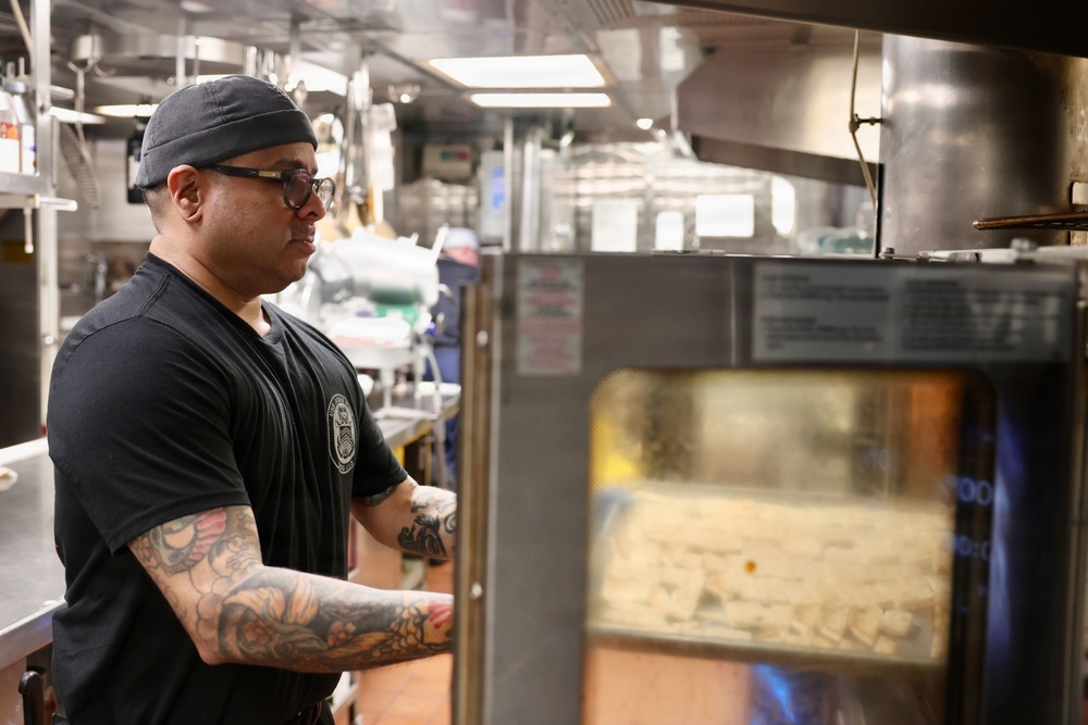A tattooed kitchen worker wearing a black t-shirt, dark cap, and glasses stands in a professional commercial kitchen, appearing to monitor or operate a large stainless steel convection oven. The foreground subject is in sharp focus while a second worker is visible in the blurred background. The setting is a busy, well-equipped restaurant or institutional kitchen.