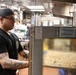 A tattooed kitchen worker wearing a black t-shirt, dark cap, and glasses stands in a professional commercial kitchen, appearing to monitor or operate a large stainless steel convection oven. The foreground subject is in sharp focus while a second worker is visible in the blurred background. The setting is a busy, well-equipped restaurant or institutional kitchen.