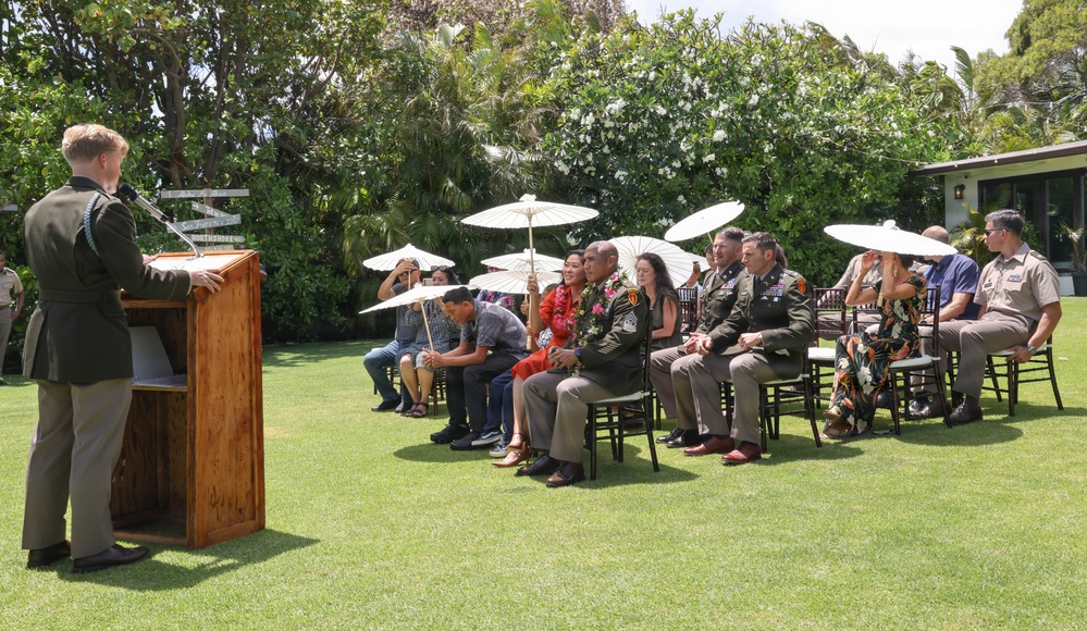An outdoor military ceremony taking place on a well-manicured lawn in what appears to be a tropical setting. A uniformed speaker addresses a seated audience that includes military personnel in Army dress uniforms and civilians, several of whom are using white parasols for sun protection. The setting suggests a formal recognition or retirement ceremony.