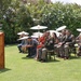 An outdoor military ceremony taking place on a well-manicured lawn in what appears to be a tropical setting. A uniformed speaker addresses a seated audience that includes military personnel in Army dress uniforms and civilians, several of whom are using white parasols for sun protection. The setting suggests a formal recognition or retirement ceremony.
