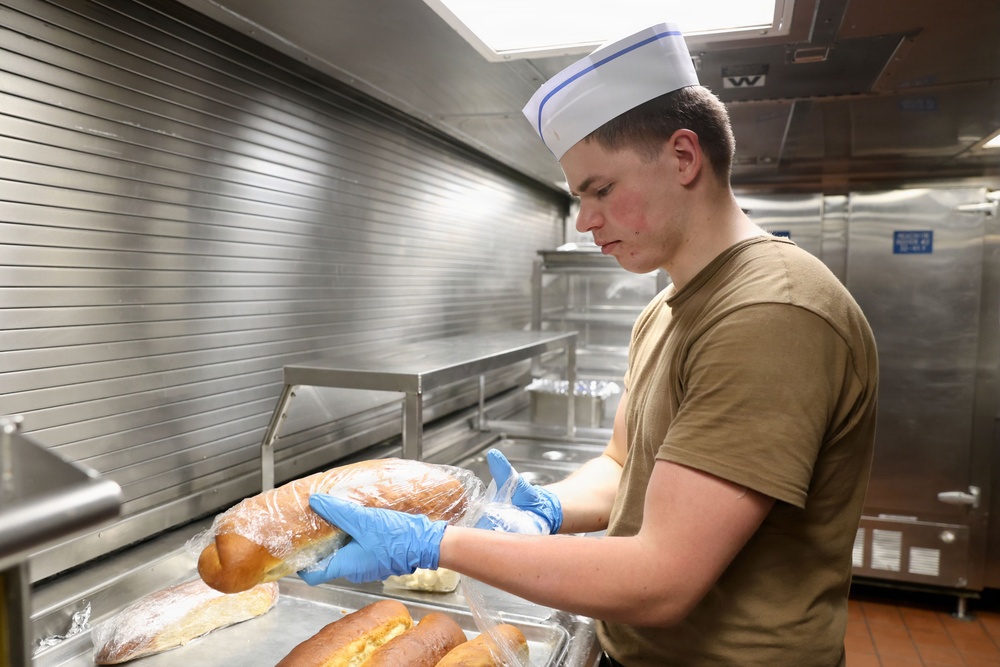 A young male food service worker wearing a white paper chef's hat and olive/tan military-style t-shirt handles wrapped bread loaves in a commercial kitchen. He is wearing blue nitrile gloves and working over a stainless steel tray containing additional bread rolls. The setting appears to be a military galley or institutional kitchen based on the stainless steel equipment and surroundings.