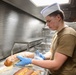 A young male food service worker wearing a white paper chef's hat and olive/tan military-style t-shirt handles wrapped bread loaves in a commercial kitchen. He is wearing blue nitrile gloves and working over a stainless steel tray containing additional bread rolls. The setting appears to be a military galley or institutional kitchen based on the stainless steel equipment and surroundings.