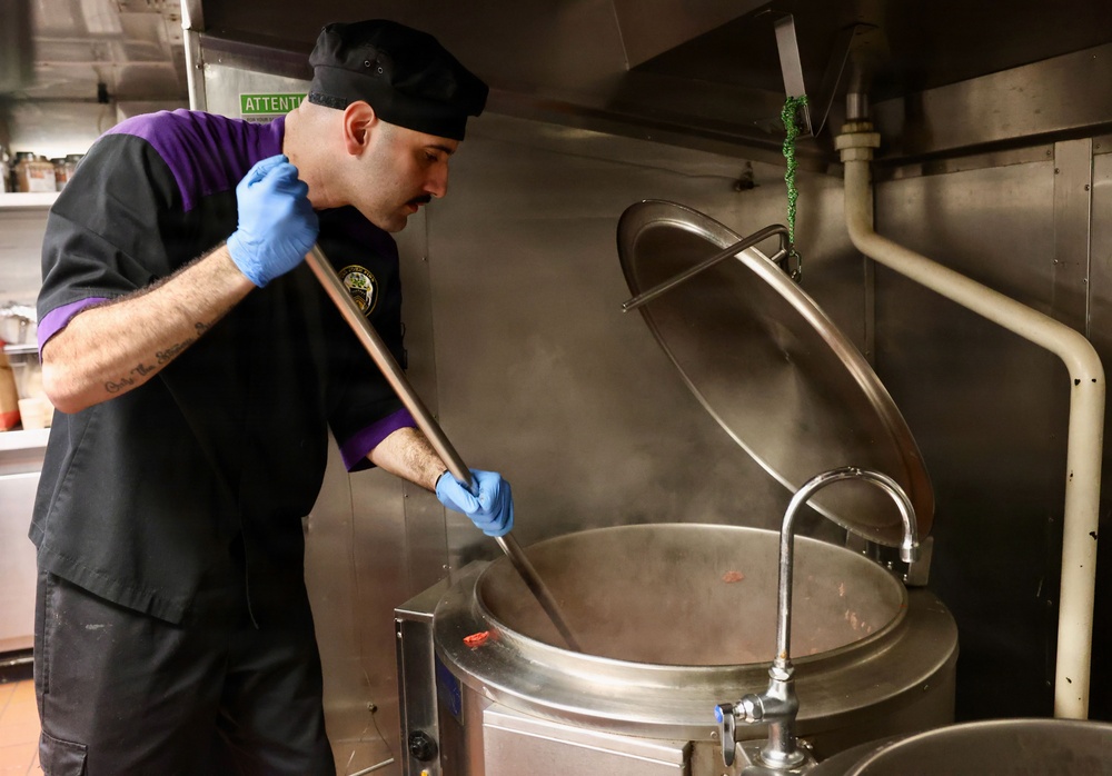 A male cook or chef is stirring a large commercial steam kettle in a professional kitchen. He is wearing a black uniform with purple accents, a black skull cap, and blue nitrile gloves. Steam is rising from the large stainless steel kettle, indicating the contents are at high temperature.
