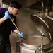A male cook or chef is stirring a large commercial steam kettle in a professional kitchen. He is wearing a black uniform with purple accents, a black skull cap, and blue nitrile gloves. Steam is rising from the large stainless steel kettle, indicating the contents are at high temperature.