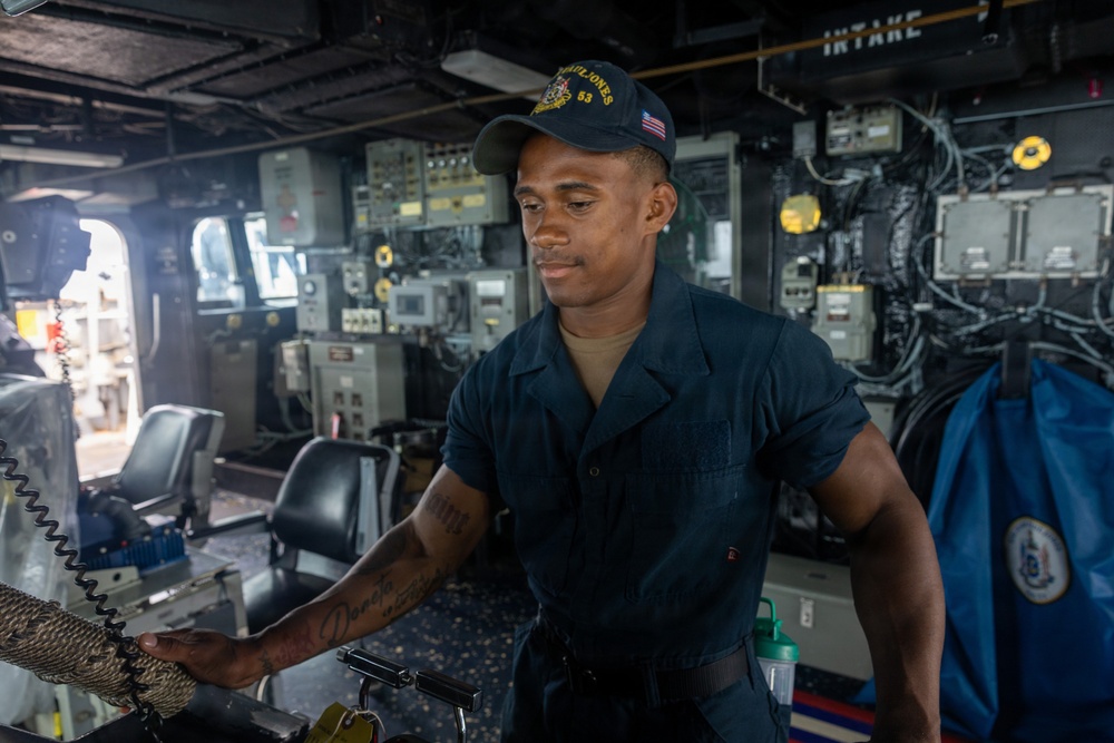 A U.S. Navy sailor is stationed at what appears to be the bridge or helm area of a naval vessel, identifiable as the USS Paul Jones (DDG-53) based on the cap insignia. The sailor is wearing a dark navy working uniform and a ship-specific ball cap with an American flag patch, and appears to be handling equipment or controls. The surrounding environment shows extensive naval electronics, control panels, and ship infrastructure.