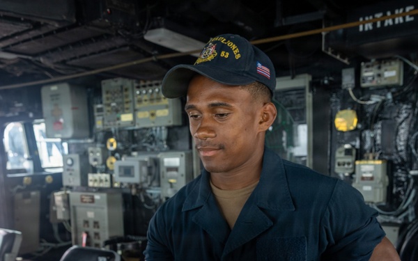 A U.S. Navy sailor is stationed at what appears to be the bridge or helm area of a naval vessel, identifiable as the USS Paul Jones (DDG-53) based on the cap insignia. The sailor is wearing a dark navy working uniform and a ship-specific ball cap with an American flag patch, and appears to be handling equipment or controls. The surrounding environment shows extensive naval electronics, control panels, and ship infrastructure.