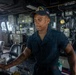 A U.S. Navy sailor is stationed at what appears to be the bridge or helm area of a naval vessel, identifiable as the USS Paul Jones (DDG-53) based on the cap insignia. The sailor is wearing a dark navy working uniform and a ship-specific ball cap with an American flag patch, and appears to be handling equipment or controls. The surrounding environment shows extensive naval electronics, control panels, and ship infrastructure.