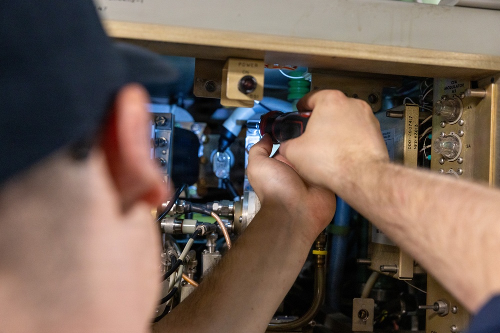 A technician wearing a dark cap is using a red-handled screwdriver to work on a complex piece of electronic or electromechanical equipment. The equipment appears to be an older, rack-mounted or cabinet-style unit with multiple circuit boards, wiring, tubes, and labeled components. The work appears to be maintenance, repair, or assembly of specialized technical hardware.