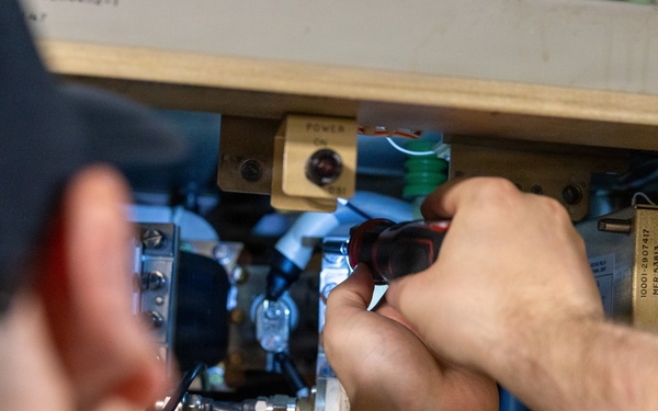 A technician wearing a dark cap is using a red-handled screwdriver to work on a complex piece of electronic or electromechanical equipment. The equipment appears to be an older, rack-mounted or cabinet-style unit with multiple circuit boards, wiring, tubes, and labeled components. The work appears to be maintenance, repair, or assembly of specialized technical hardware.